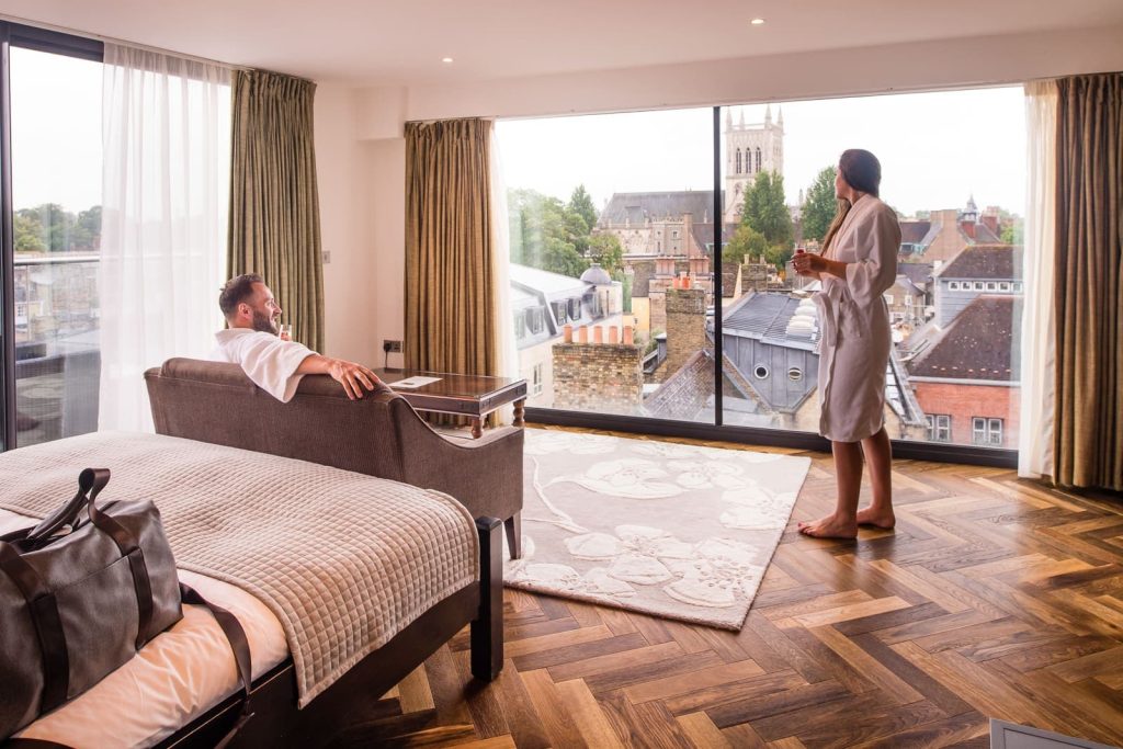 couple in hotel bedroom, both wearing dressing gowns and drinking champagne looking at the view of cambridge