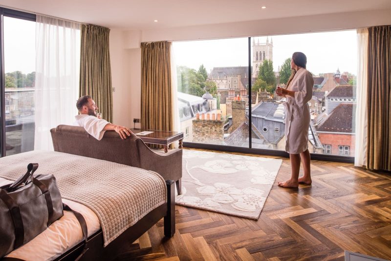 couple in hotel bedroom, both wearing dressing gowns and drinking champagne looking at the view of cambridge