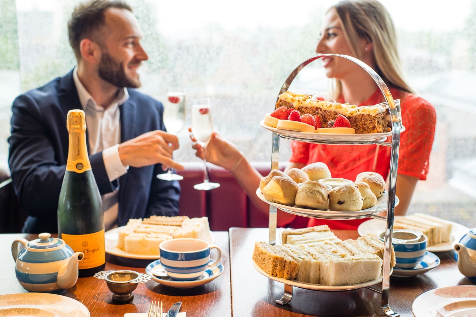 a couple clinking champagne sitting behind their afternoon tea of sandwiches, scones with jam and cream and sweet treats