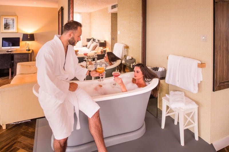 couple in hotel bathroom with woman in roll top bath holding a cocktail cheering her partner who is sitting on the edge