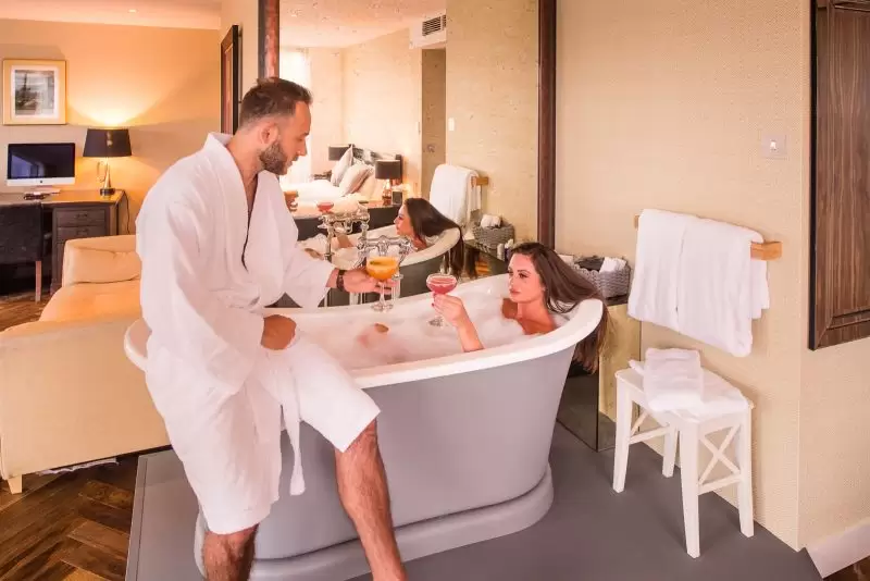 couple in hotel bathroom with woman in roll top bath holding a cocktail cheering her partner who is sitting on the edge