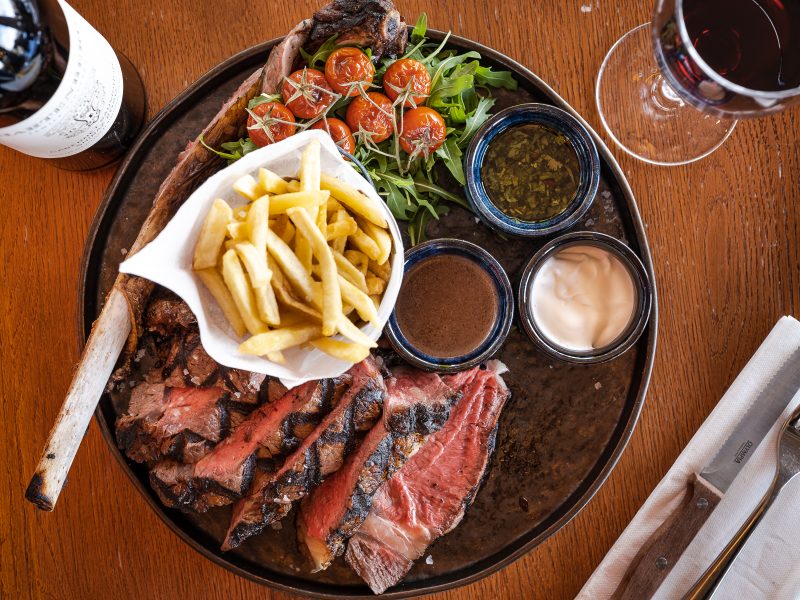 a round plate of t-bone steak with different sauces and a bucket of chips, with red wine in a glass