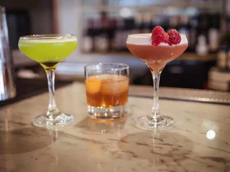 three cocktails on the bar, two cosmopolitan type drinks one acid green another is pink with raspberries, and a short drink looks like whiskey
